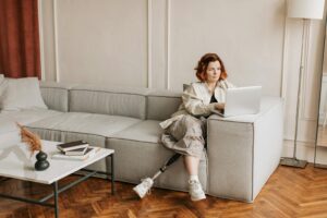 A woman with a prosthetic leg works remotely on a laptop in a cozy living room setting.