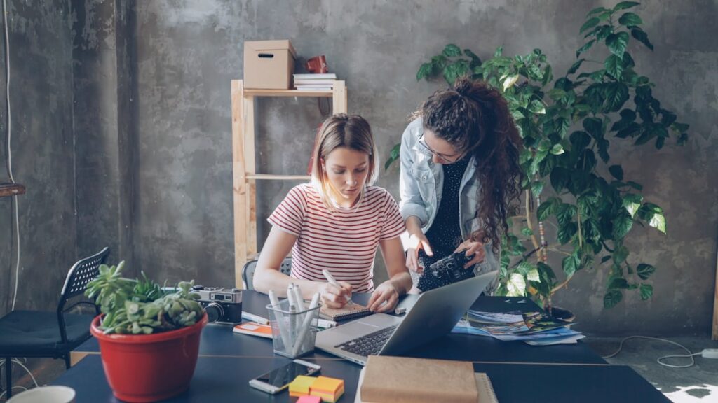 Young designer is drawing images in notebook sitting at table while female photographer is coming to her with camera. Women start discussing new project and watching photos on screen.
