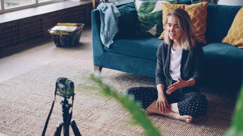 Pretty blond woman is talking and looking at camera recording video for her online blog sitting on floor in modern flat. Technology, mass media and creative people concept.