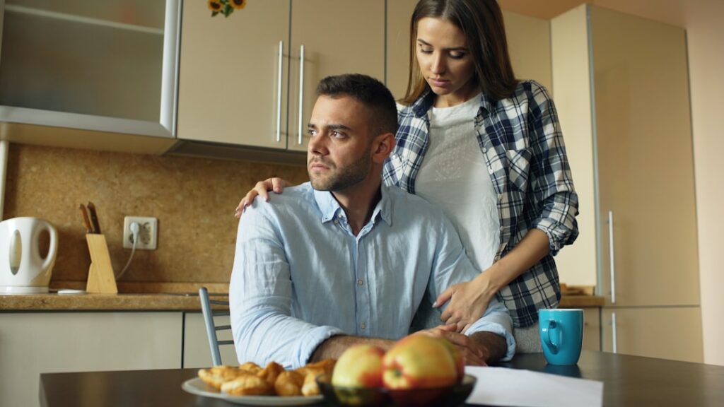 Upset young man reading unpaid bills and hugged by his wife supporting him early morning in the kitchen at home