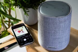 This image showcases a sleek Harman Kardon smart speaker positioned next to a smartphone on a wooden surface. The scene highlights the seamless integration of smart home technology, where voice-activated devices and mobile apps work together to deliver high-quality audio experiences. The minimalist design of the speaker, paired with natural elements like a potted plant, creates a cozy and modern atmosphere perfect for any smart home setup. Ideal for illustrating themes around smart assistants, home automation, and wireless music streaming.