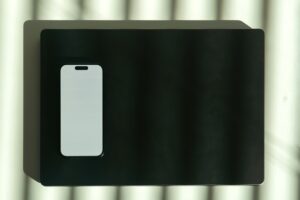Smartphone mockup screen Laptop Shadows Minimalist Workspace Top-down view of a modern smartphone placed on a closed laptop, with striped shadows creating a dramatic lighting effect. Clean and minimal composition suitable for technology, remote work or digital lifestyle concepts