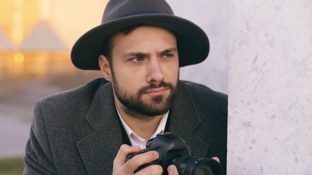 Closeup of Young paparazzi man in hat photographing celebrities on camera while spy behind wall