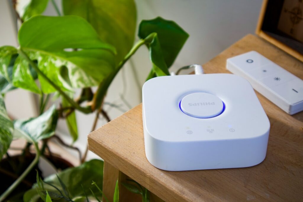 This image showcases a Philips Hue Bridge, the central hub for Philips Hue smart lighting systems, resting on a wooden table beside a minimalist remote control. The scene is brightened by the presence of vibrant green houseplants, suggesting a harmonious blend of technology and nature. The glowing blue ring on the bridge indicates it is powered and connected, ready to control a network of smart bulbs and lights throughout the home. Ideal for illustrating smart home automation, modern living, or energy-efficient lighting solutions, this image conveys a sense of convenience and innovation.